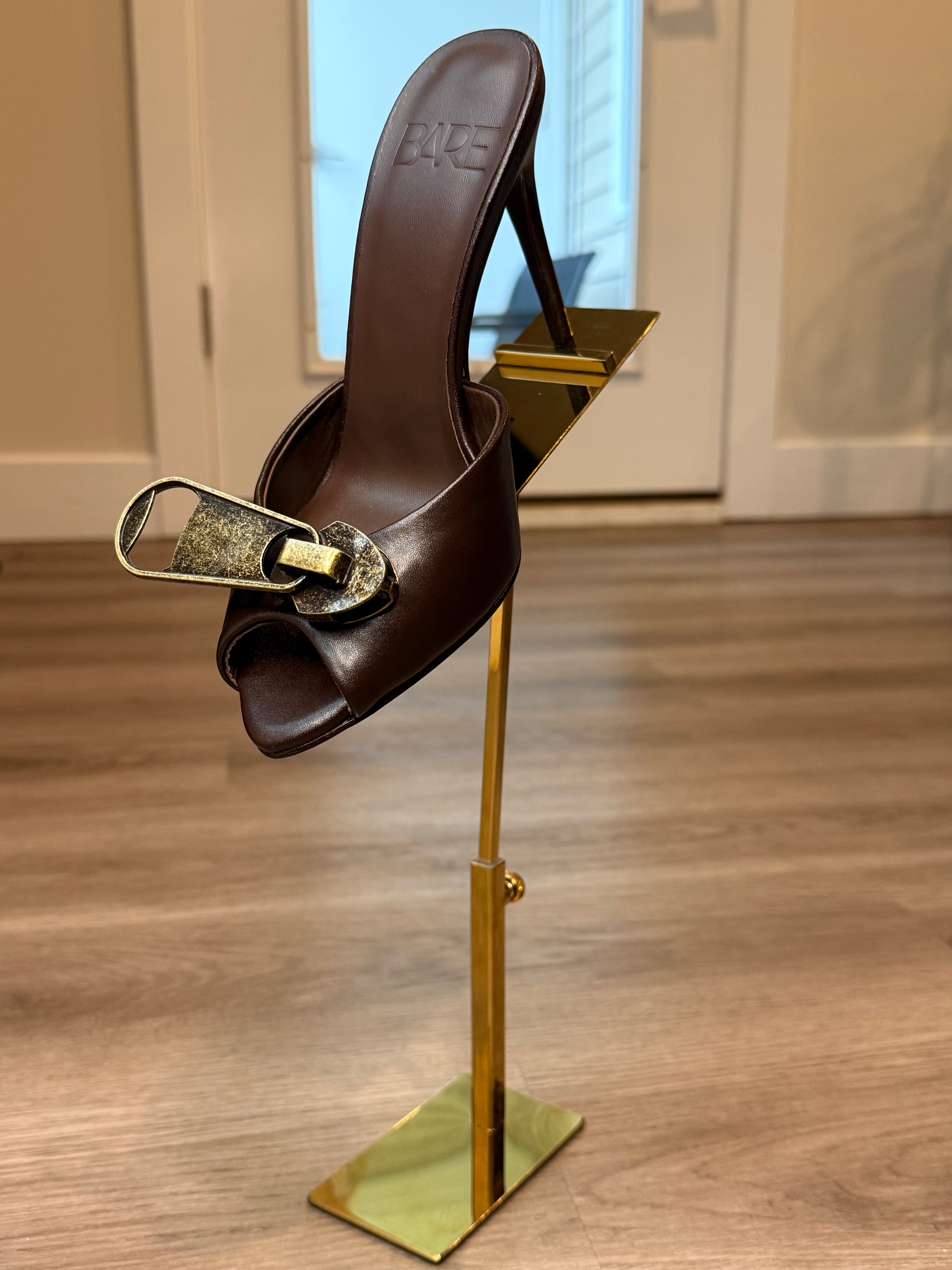 Brown sandal with gold buckle on a gold stand against a neutral background