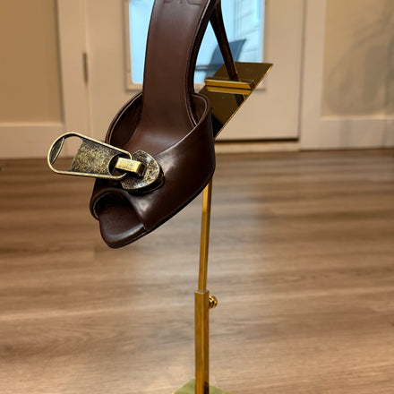 Brown sandal with gold buckle on a gold stand against a neutral background